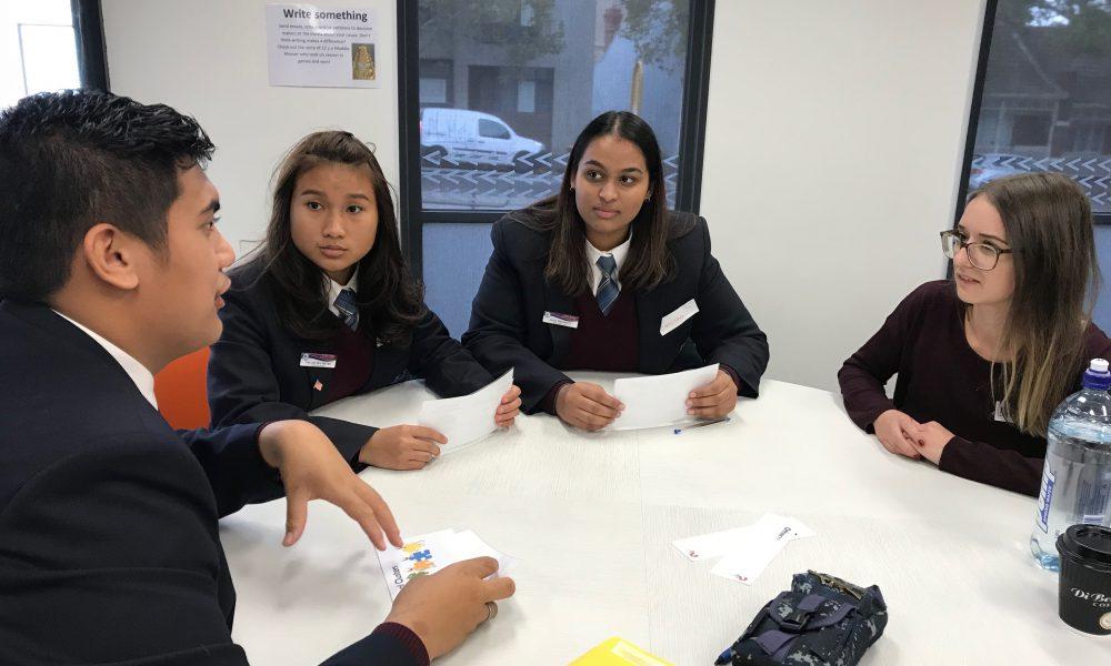 A group of students sit around a table, talking to a teacher.