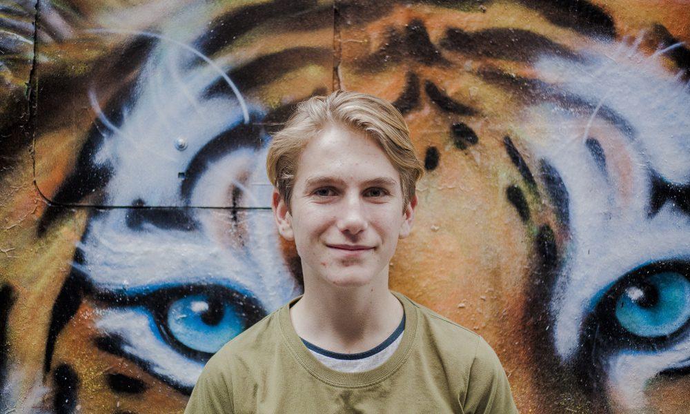 The author of the piece smiles at the camera as he stands in front of a mural.