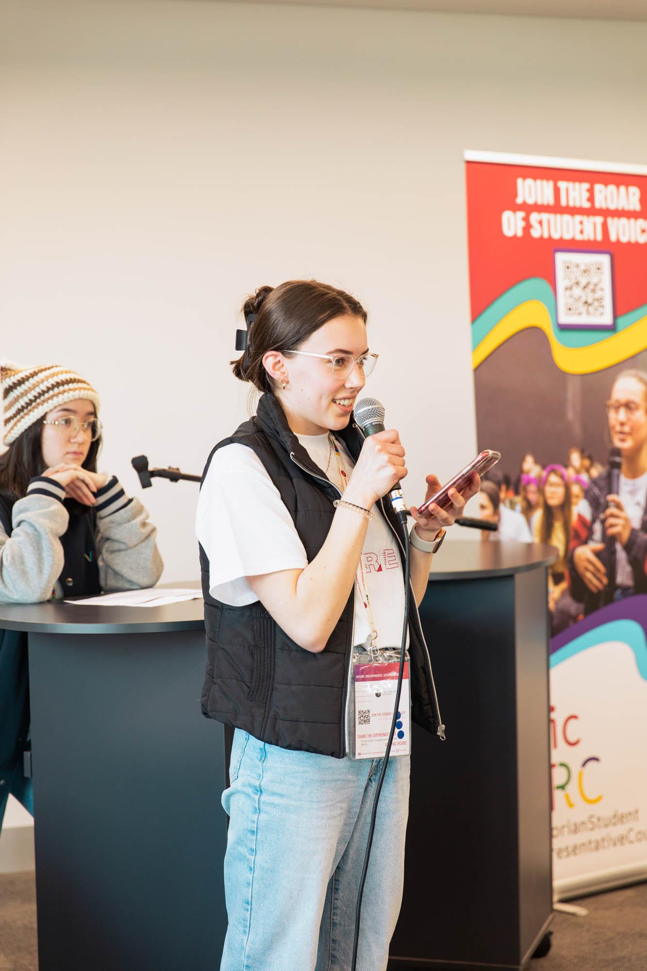 Arwen stands with a microphone at VicSRC Congress 2022, pitching her issue in education.