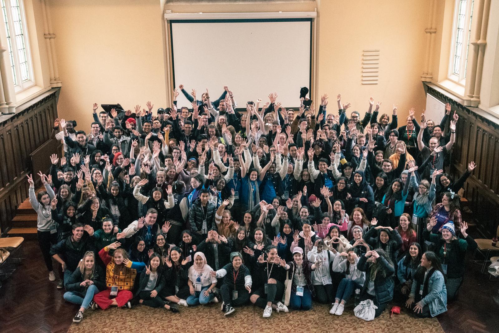 A big group of students with their arms in the air in celebration all smile at the camera.