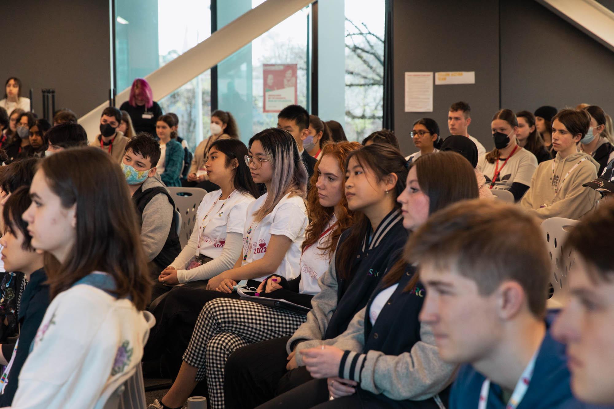 A group of students sit on chairs arranged in rows listening intently.