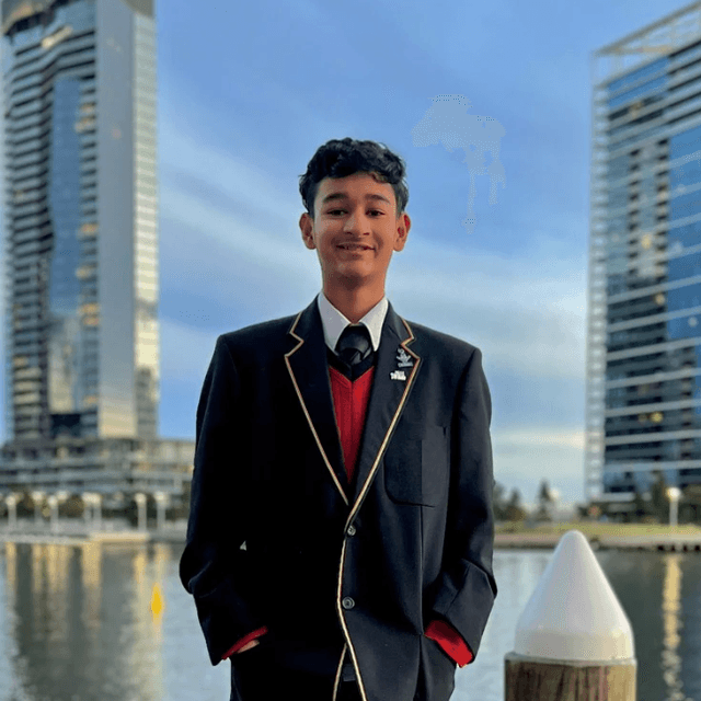 A photo of a boy posing in a suit in front of a body of water and some skyscrapers.