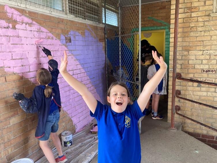 Primary school student puts their hands in the air with excitement, while another paints the wall behind them.