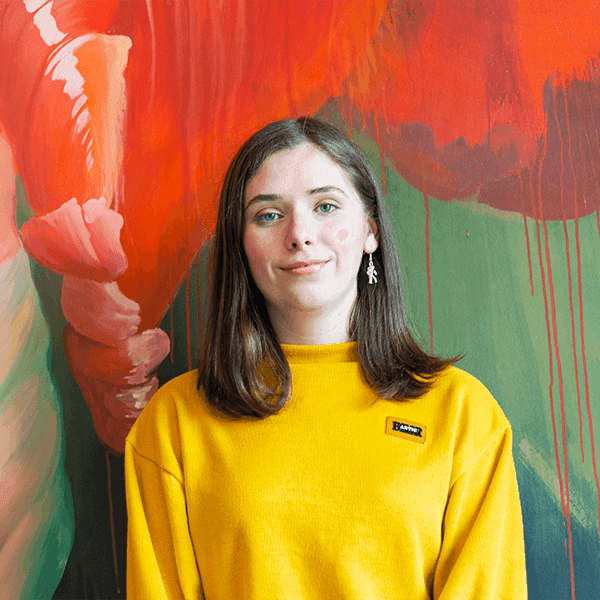 A young woman with light brown hair in a yellow cardigan stands against a predominantly red artwork