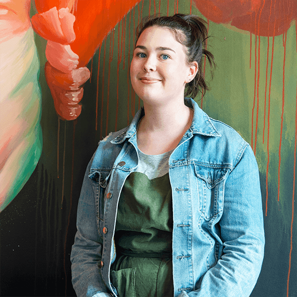 Emily, a young woman with pulled back brown hair wearing a green blouse and denim jacket stands in front of a painting.